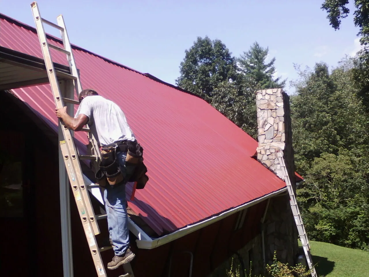 Roofer on a ladder beside a red metal roof and stone chimney — Soffit & Fascia Repair by Roofing Brothers in Vandergrift